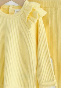 Pulover galben din tricot cu model în relief și detaliu cu volane pe umăr, asortat cu pantaloni din tricot cu model în relief. Textură moale și croială confortabilă.