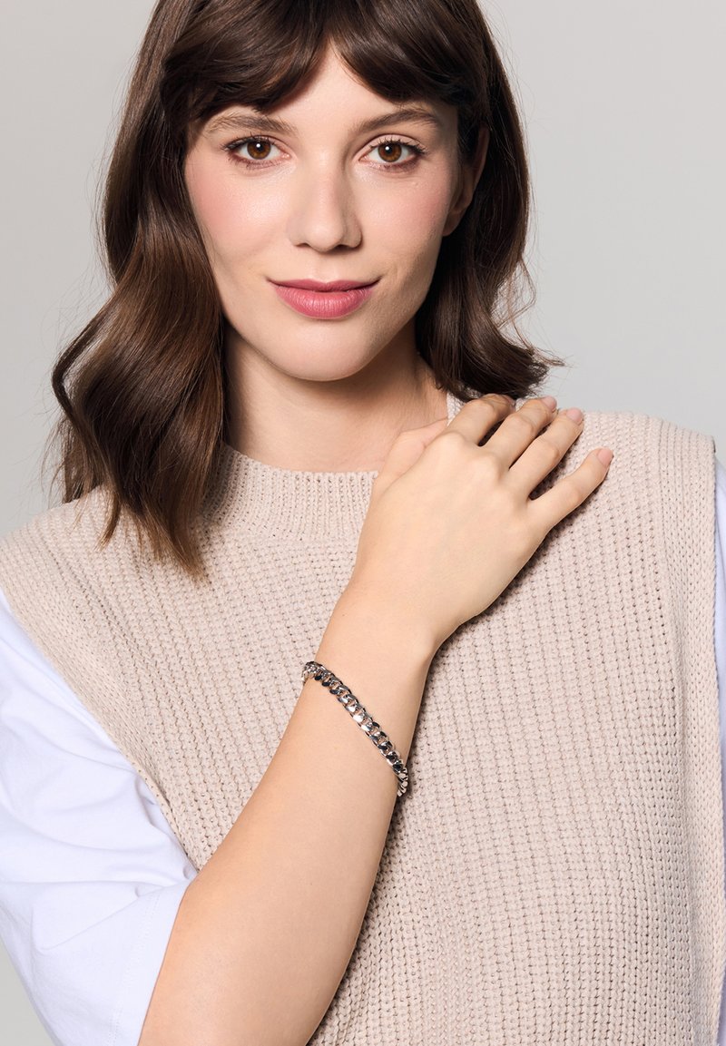 Srebrna bransoletka łańcuszkowa noszona na nadgarstku, z splatającymi się ogniwami. Tło jest neutralne, podkreślając wzór i teksturę bransoletki.