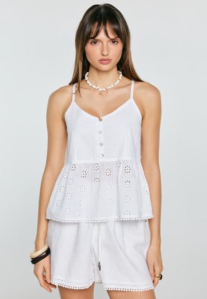 Kobieta w białym bezrękawnym topie ażurowym z guzikami i pasujących spodenkach, z dodatkami w postaci naszyjnika z muszli i bransoletek, na jasnym tle.