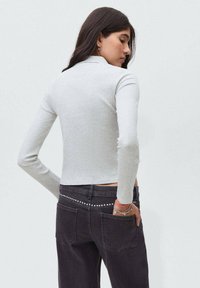 Kvinna med långt mörkt hår iklädd en ljusgrå ribbad långärmad tröja och mörka jeans med ett nitarbälte, vänd åt sidan med handen i fickan.