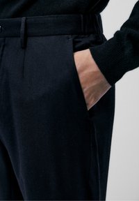 Zwarte, op maat gemaakte broek met een gladde textuur, voorzien van zijzakken en een strakke pasvorm. Riemlussen zichtbaar aan de taille.