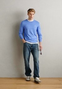 Hellblauer Strickpullover über einem weißen Hemd, kombiniert mit blauen Jeans und grauen Streifenschuhen. Trägt dunkle Sonnenbrillen.