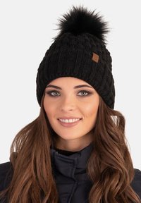 Bonnet en tricot noir avec un motif texturé et un pompon en fausse fourrure sur le dessus. Comprend une petite étiquette en cuir marron sur le côté.