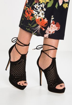 Czarne sandały na wysokim obcasie z siateczkowym wzorem, otwartym noskiem i paskami na kostkę. Zamszowa faktura i falujące krawędzie podkreślają styl.