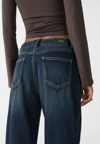 Tmavě modré džíny z denimu s vysokým pasem, obsahující dvě zadní kapsy a štítek na pase. Materiál má mírnou strukturu.