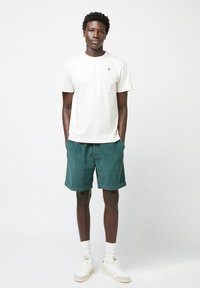 T-shirt bianco in cotone con un piccolo logo di un albero verde, abbinato a pantaloni corti in velluto a coste verde scuro. Le scarpe sono bianche con dettagli verde chiaro.