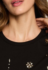 Sort stof t-shirt med rund halsudskæring, prydet med dekorative udsmykninger i guld og klare krystalformer, der skaber kontrasterende tekstur og detaljer.
