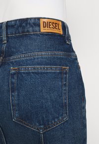 Mörkblå denimjeans med en stor brun läderlapp märkt "DIESEL" på bakre midjan. Har orange sömmar och en bakficka.