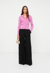 Frau in einem rosa, geknöpften Cardigan und schwarzen, weiten Cargo-Hosen, die eine kleine, beigefarbene Handtasche hält.