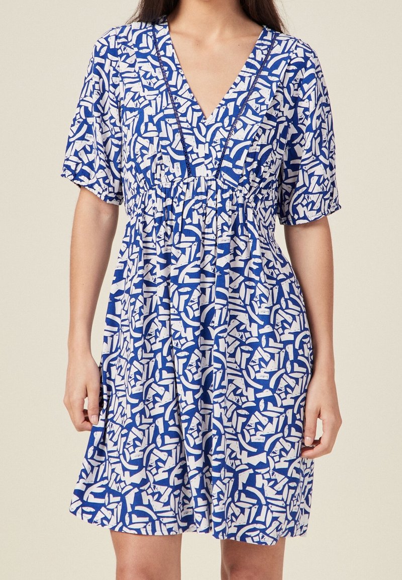 Femme portant une robe à imprimé abstrait bleu et blanc avec des manches courtes bouffantes et un décolleté en V, debout sur un fond uni.