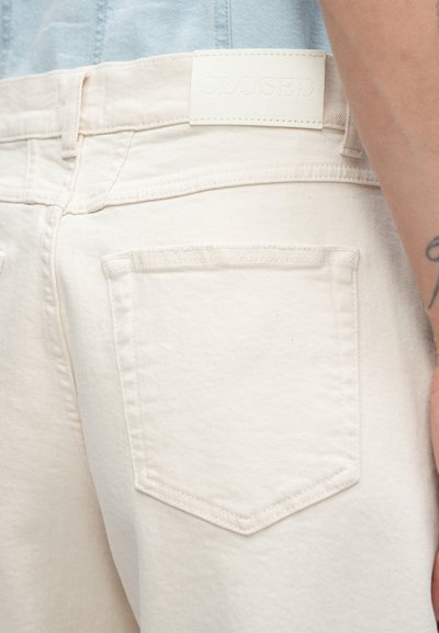 Jean en denim beige avec une taille mi-haute, deux poches arrière et une étiquette en cuir blanc texturé avec le logo "CLOSED" embossé.