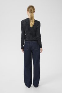 Egy szőke lófarokkal rendelkező nő fekete bordás pulóvert és sötétkék, bő szárú farmernadrágot visel, háttal áll egy sima háttér előtt.