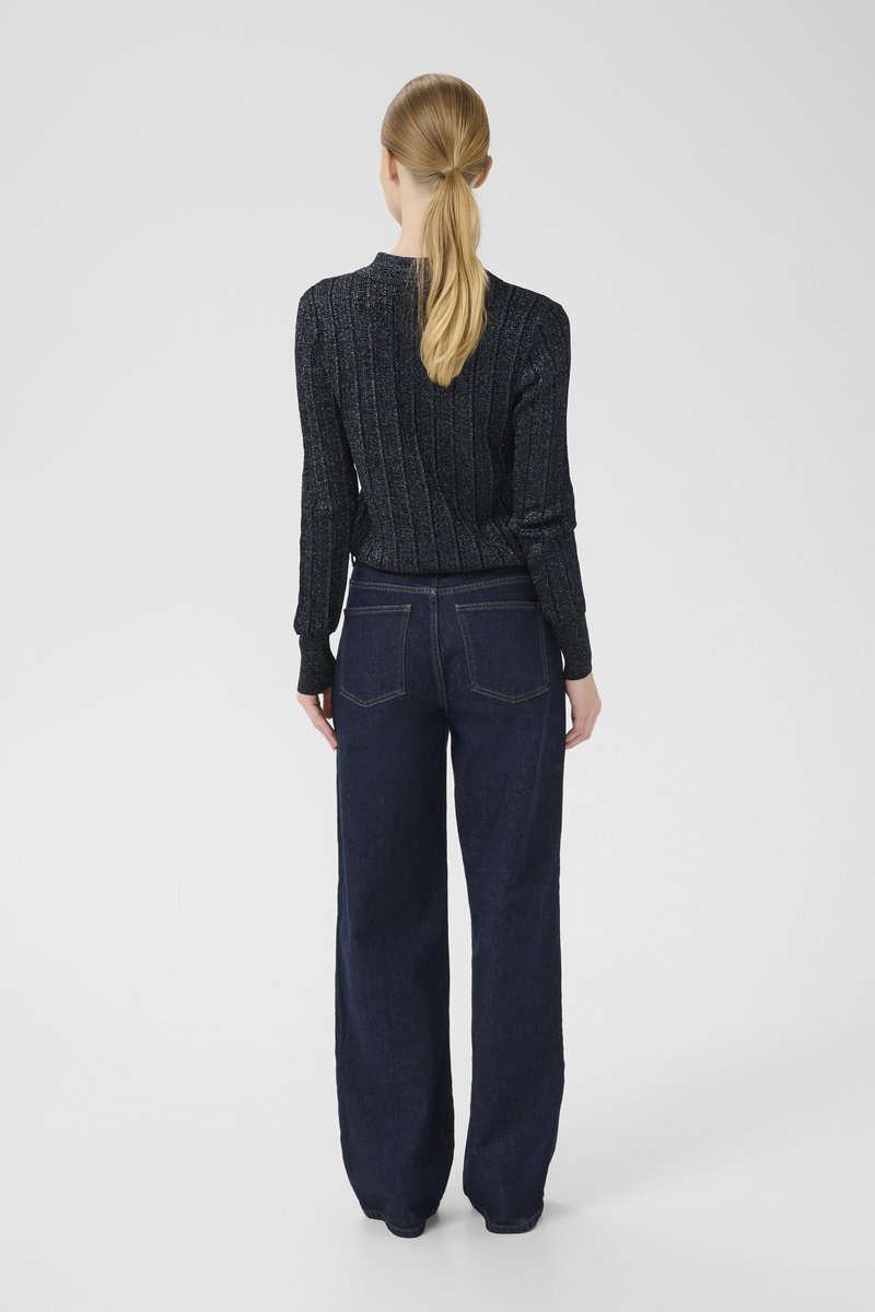 Frau mit blonden Pferdeschwanz, die einen schwarzen gerippten Pullover und dunkle, weit geschnittene Jeans trägt, steht mit dem Rücken zu einem einfarbigen Hintergrund.