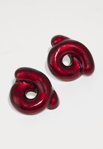 Κόκκινα γυαλιστερά σκουλαρίκια σε σχήμα σπείρας με απαλή υφή και ανοιχτό κέντρο, διαθέτοντας μια γλυπτική σχεδίαση που τονίζει τις καμπύλες.