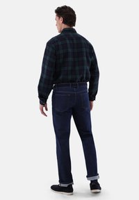 Camicia a quadratini verde e blu navy, jeans blu scuro, arrotolati alla caviglia, e scarpe nere, viste da dietro.