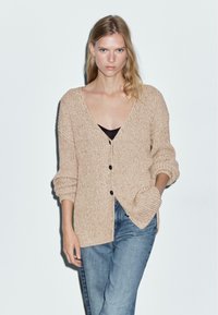 Beige stickad cardigan med V-ringning, långa ärmar och textilstruktur. Har två framfickor och tre svarta knappar. Bärs med jeans.
