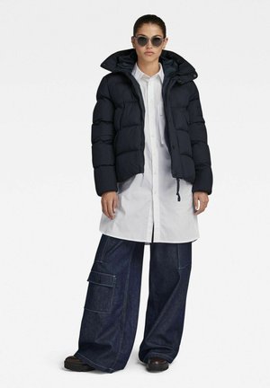 Woman wearing dark puffer jacket, long white buttoned shirt, wide-leg dark denim pants with pocket, brown shoes, and round sunglasses, standing.