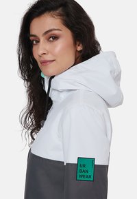 Balta ir pilka su gobtuvu striukė su juodu užtrauktuku, turinti žalią logotipo ekrano užsklandą ant rankovės ir raištelį gobtuve. Lygus audinys.