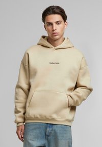 Beige hoodie met een voortasje, geribde manchetten en een bedrukte logo "AnotherCotton" in het midden. Zacht katoenmix materiaal.