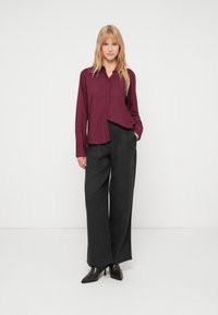Bordeaux-langärmliges Button-Down-Hemd, lockere Passform, kombiniert mit grauen karierte High-Waist-Hosen mit weitem Bein und spitzen schwarzen High Heels.