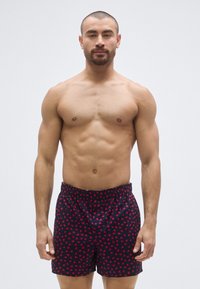 Tengerészkék fürdőnadrág piros szív mintával, rugalmas derékrésszel és egyenes szabással. A modell mezítelen felsőtesttel áll, bemutatva a dizájnt.