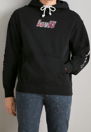 Osoba nosząca czarną bluzę z kapturem Levi’s z kolorowym logo, białym ściągaczem i napisem na prawym rękawie, w połączeniu z niebieskimi jeansami acid-washed.