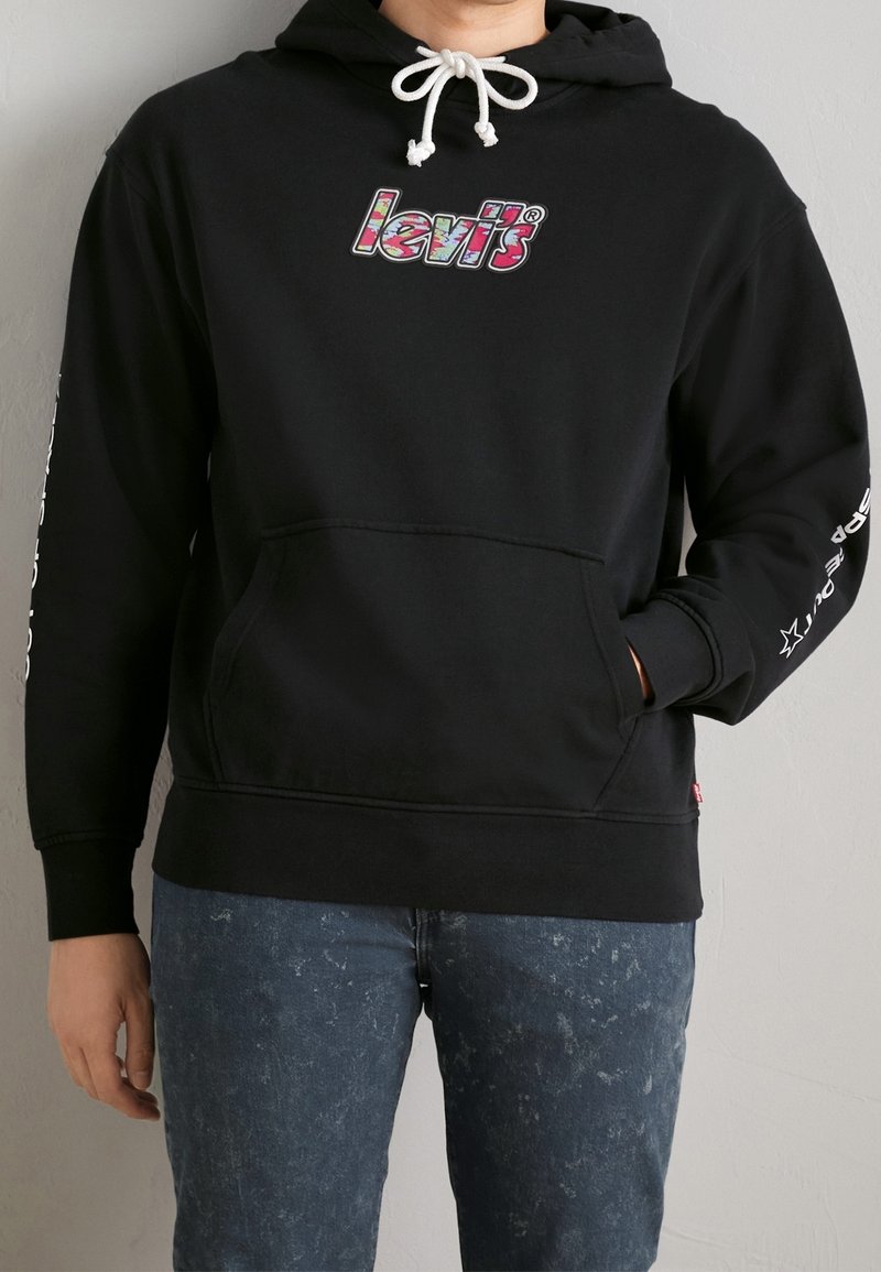 Personne portant un sweat à capuche noir Levi’s avec un logo coloré, un cordon blanc et un texte sur la manche droite, associé à un jean bleu délavé à l'acide.