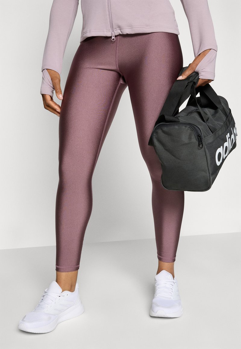 Lila, figurbetonte Leggings mit glatter Textur; kombiniert mit einer schwarzen Sporttasche mit weißer Markenbeschriftung. Weiße Sportschuhe runden den Look ab.