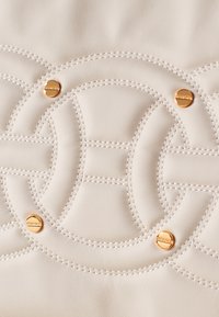 Cremfarbige Lederhandtasche mit komplizierten Nähten in einem kreisförmigen Muster, verziert mit kleinen goldenen Hardware-Elementen. Glatte Textur.