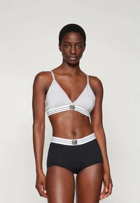 Siva bralette z tanjšimi naramnicami in globokim V-izrezom, v kombinaciji s črnimi kratkimi hlačami z visokim pasom, ki imajo bele črte in okrasni logo.