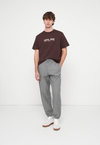 Rudas medvilninis marškinėlis su baltu logotipo užrašu, derinamas su šviesiai pilkomis kelnėmis su raišteliu ir baltais sportbačiais su rudais padais.