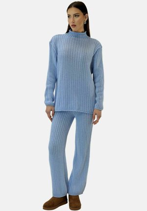 Donna con maglione a coste azzurro chiaro e pantaloni abbinati, abbinati a stivaletti marroni, in piedi davanti a uno sfondo semplice.