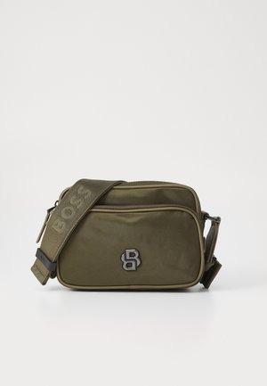 Oliwkowa torba na ramię z regulowanym paskiem, z wyraźnym czarnym logo i zapięciem na zamek. Prostokątny kształt o gładkiej teksturze.