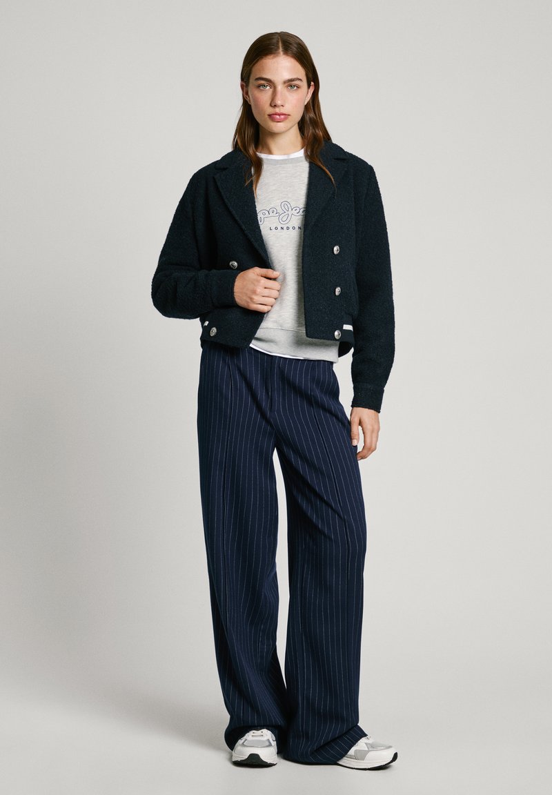 Dunkelmarinefarbige, kurz geschnittene Jacke mit silbernen Knöpfen, grauer Sweatshirt mit Logo, vertikal gestreifte weite Hose und weiße Sportschuhe.