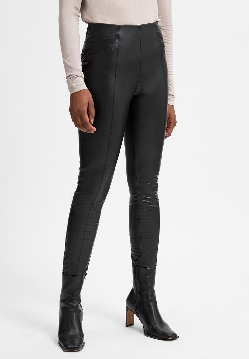 Leggings en simili cuir noir taille haute avec un design épuré affichant des coutures latérales et une texture lisse, associés à des bottes à talons noires.