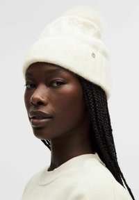 Kreemi värvi kudumine beanie, mille peal on koheline pom-pom, ribakujundusega ja küljes väike dekoratiivne metallist aksessuaar.