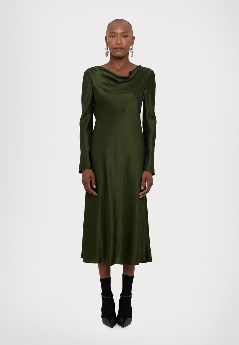 Langsleeve, olijfgroene jurk met een cowl-halslijn, gemaakt van gladde stof, met een vloeiende rok, gecombineerd met zwarte hakken en sokken.