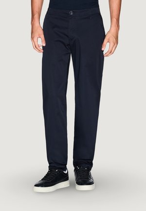 Pantaloni blu navy in tessuto liscio, con chiusura a bottone, tasche laterali e design a gamba dritta. Abbinati a sneakers nere.
