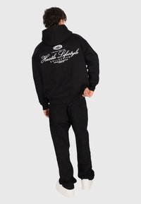 Person, der einen schwarzen Hoodie und eine Hose trägt, mit weißem Text "Hustle Lifestyle Collection" auf dem Rücken, steht vor neutralem Hintergrund.
