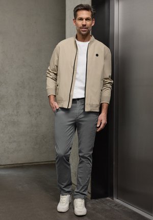 Mies seisoo sisätiloissa ja hänellä on yllä beige takki, valkoinen t-paita, harmaat housut ja valkoiset tennarit. Hän nojaa kevyesti tummaa seinää vasten hissin lähellä.