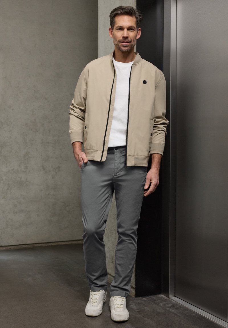 Homme debout à l'intérieur portant une veste beige, un t-shirt blanc, un pantalon gris et des baskets blanches, s'appuyant légèrement contre un mur sombre près de l'ascenseur.