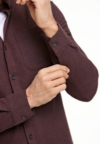 Burgundin nappipaita teksturoidusta kankaasta, jossa on rintatasku ja mustat napit. Kuvassa on lähikuva kädestä, joka säätää hihansuut.