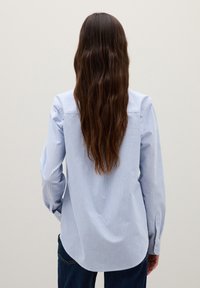 Persona con capelli lunghi castani che indossa una camicia a righe verticali celeste e bianca e jeans blu scuro, in piedi di fronte a uno sfondo neutro.