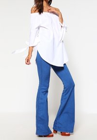 Donna che indossa una blusa bianca scollata con maniche legate, jeans a zampa d'elefante blu e sandali con piattaforma marroni, in piedi davanti a uno sfondo semplice.