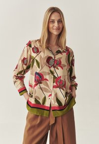 Jonge vrouw met een beige bloemetjesoverhemd met rode, groene en zwarte accenten, gecombineerd met losse bruine broeken, handen in de zakken.
