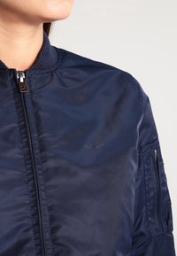 Námorně modrá bomber bunda vyrobená z hladkého nylonu. Má přední zip, žebrovaný límec a kapsu na zip na levém rukávu.