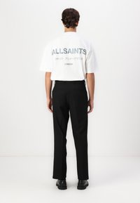 Λευκό κοντομάνικο μπλουζάκι με εκτύπωση "ALLSAINTS" στην πλάτη, συνδυασμένο με μαύρο παντελόνι ίσιας γραμμής και μαύρα παπούτσια, μπροστά σε απλό φόντο.