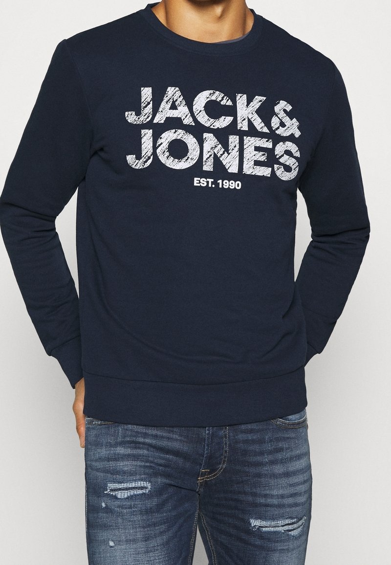 Man som bär en marinblå sweatshirt med vit text "JACK & JONES EST. 1990" och slitna blåjeans, med händerna i fickorna.