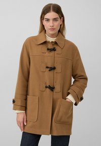 Frau in einem beigen Toggelmantel mit großen Fronttaschen, schwarzen Verschlüssen und umgeschlagenen Ärmeln über einem cremefarbenen Strickpullover und dunklen Hosen.