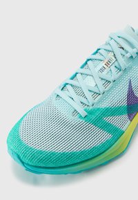 Le scarpe da ginnastica di colore azzurro chiaro presentano una tomaia in rete con accenti verdi e viola, lacci piatti e una suola a gradiente che transita dal teal al giallo.